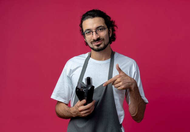 Blij jonge blanke mannelijke kapper bril en golvende haarband dragen uniform houden en wijzend op tondeuses, kam, spray fles geïsoleerd op crimson achtergrond met kopie ruimte