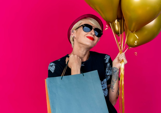 Gratis foto blij jonge blonde feestvrouw met feestmuts en zonnebril met ballonnen en papieren zakken geïsoleerd op roze muur met kopie ruimte