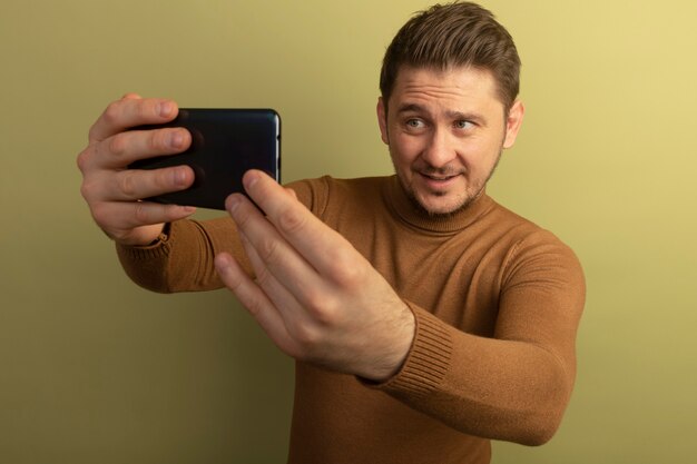 Blij jonge blonde knappe man selfie te nemen geïsoleerd op olijfgroene muur