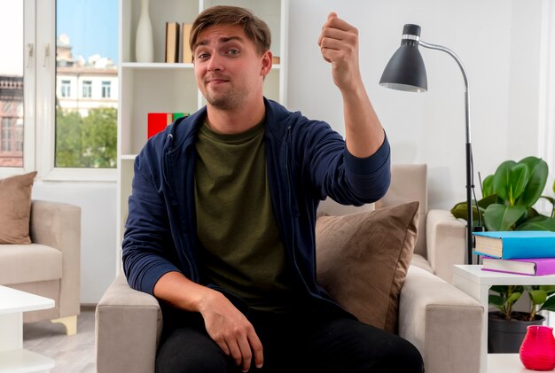 Blij jonge blonde knappe man zit op fauteuil houden vuist kijken camera in de woonkamer