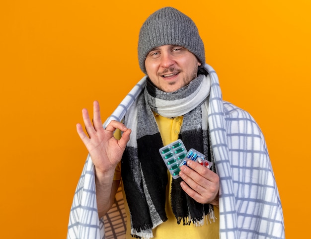 Blij jonge blonde ziek Slavische man met winter muts en sjaal gebaren oke hand teken en houdt pakje medische pillen verpakt in plaid geïsoleerd op oranje muur met kopie ruimte