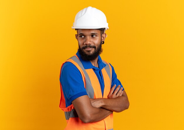 blij jonge bouwer man in uniform met veiligheidshelm staande met gekruiste armen geïsoleerd op oranje muur met kopieerruimte