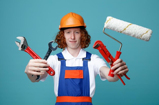 Blij jonge bouwer man in uniform stak bouwgereedschap op camera geïsoleerd op blauwe achtergrond