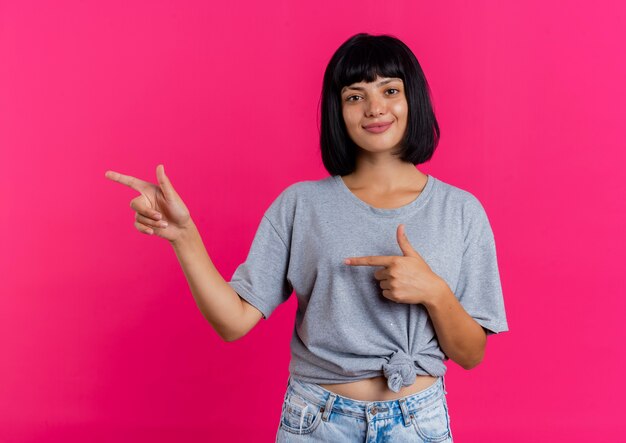 Blij jonge brunette blanke meisje wijst naar kant met twee handen kijken camera geïsoleerd op roze achtergrond met kopie ruimte