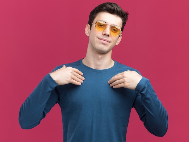 Blij jonge brunette kaukasische jongen in zonnebril legt handen op de borst camera kijken op roze