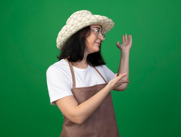 Blij jonge brunette vrouwelijke tuinman in optische bril en uniform dragen tuinieren hoed wijst terug met handen geïsoleerd op groene muur