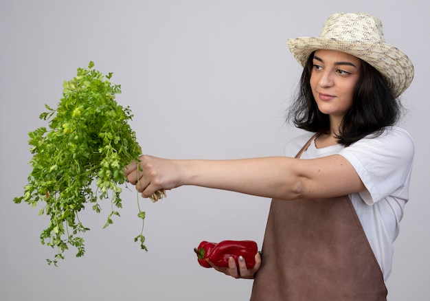 Blij jonge brunette vrouwelijke tuinman in uniform dragen tuinieren hoed houdt rode paprika's en kijkt naar koriander geïsoleerd op een witte muur met kopie ruimte