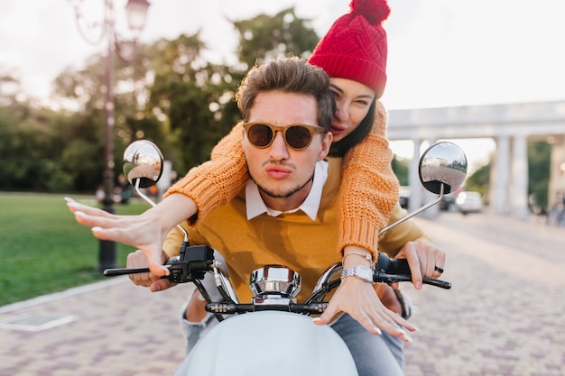 Blij jonge dame in trendy accessoires en gebreide trui genieten van snelle rit met vriendje