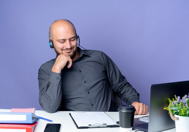 Blij jonge kale call center man met hoofdtelefoon achter bureau met werk tools kijken naar Klembord met hand op kin geïsoleerd op paarse achtergrond