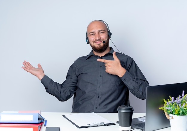 Blij jonge kale call center man met hoofdtelefoon zittend aan een bureau met uitrustingsstukken met lege hand en wijzend op het geïsoleerd op een witte achtergrond