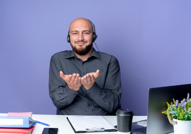 Blij jonge kale call center man met hoofdtelefoon zittend aan een bureau met uitrustingsstukken met lege handen op camera geïsoleerd op paarse achtergrond