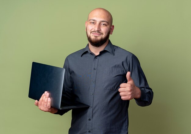 Blij jonge kale call center man met laptop en duim opdagen geïsoleerd op olijfgroene achtergrond met kopie ruimte