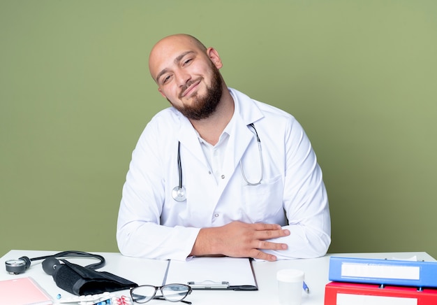 Blij jonge kale mannelijke arts die medische mantel en stethoscoop zittend aan een bureau werkt met medische hulpmiddelen geïsoleerd op een groene achtergrond