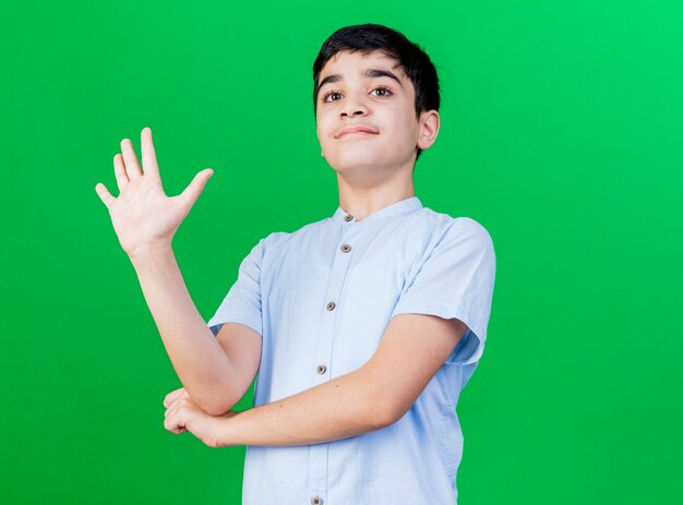 Blij jonge Kaukasische jongen die camera bekijkt die hand onder elleboog houdt die vijf met hand toont die op groene achtergrond met exemplaarruimte wordt geïsoleerd