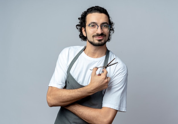 Blij jonge Kaukasische mannelijke kapper bril en golvende haarband dragen uniform staande met gesloten houding en houden schaar geïsoleerd op een witte achtergrond met kopie ruimte