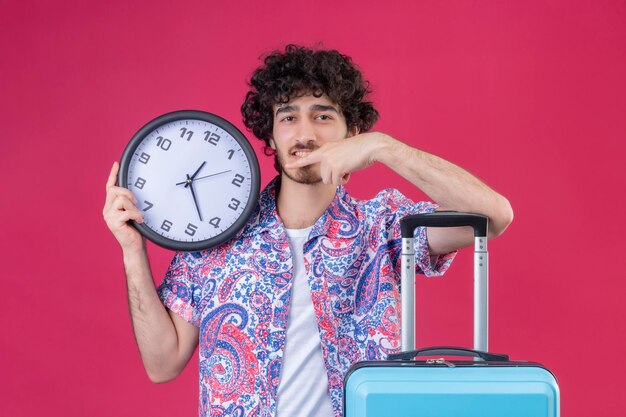 Blij jonge knappe gekrulde reiziger man met klok erop wijzend en arm op koffer op geïsoleerde roze ruimte