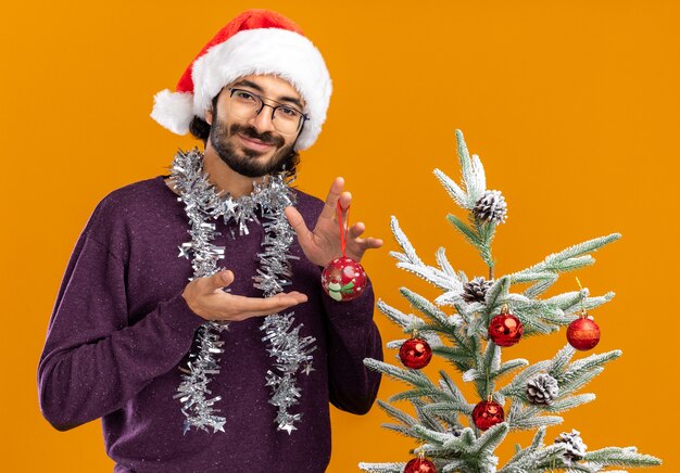 Blij jonge knappe jongen permanent in de buurt van kerstboom met kerstmuts met slinger op nek houden en wijst naar kerstballen geïsoleerd op een oranje achtergrond