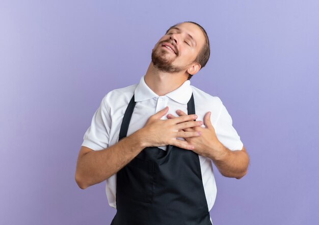 Blij jonge knappe kapper die uniform draagt ?? handen op zijn hart met gesloten ogen geïsoleerd op paarse muur