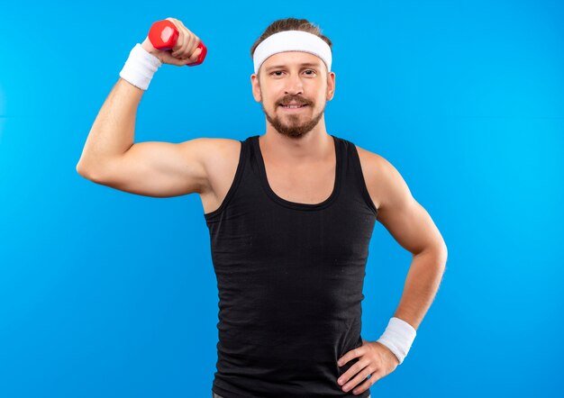 Blij jonge knappe sportieve man met hoofdband en polsbandjes halter opheffen en hand op taille zetten geïsoleerd op blauwe ruimte