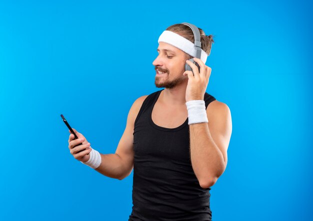 Blij jonge knappe sportieve man met hoofdband en polsbandjes met koptelefoon houden en kijken naar mobiele telefoon met één hand op hoofdtelefoon geïsoleerd op blauwe ruimte
