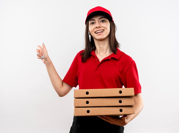 Blij jonge levering meisje dragen rode uniform en pet houden stapel pizzadozen wijzend met wijsvinger naar de zijkanten glimlachend vrolijk staande op witte achtergrond