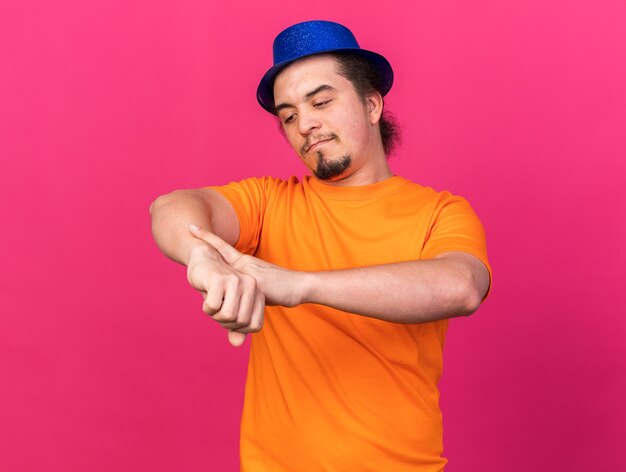 Blij jonge man met feestmuts en het tonen van pols klok gebaar geïsoleerd op roze muur