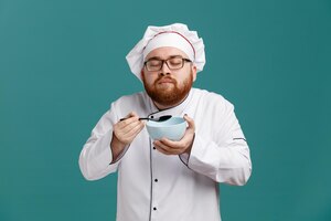 Blij jonge mannelijke chef-kok dragen glazen uniform en dop met lepel en lege kom doen alsof ruikende geur van voedsel met gesloten ogen geïsoleerd op blauwe achtergrond