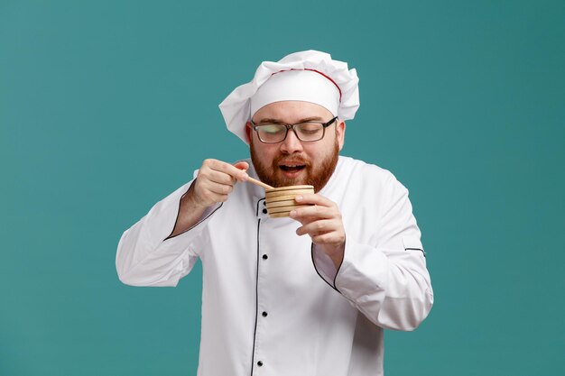 Blij jonge mannelijke chef-kok met bril uniform en pet met houten kom met honingdipper erin in de buurt van mond grijpen honinglepel ruikende geur van honing met gesloten ogen