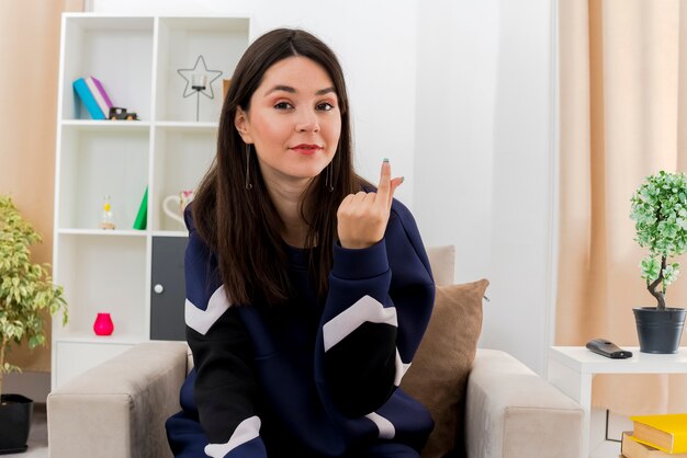 Blij jonge mooie blanke vrouw zittend op een fauteuil in ontworpen woonkamer op zoek en geld gebaar doen