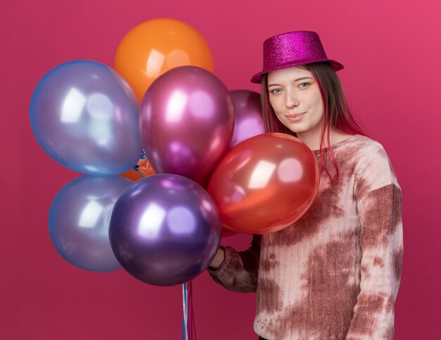 Blij jonge mooie dragen feestmuts met ballonnen geïsoleerd op roze muur