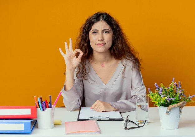 Blij jonge mooie vrouwelijke beambte zittend aan een bureau met office-hulpprogramma's die ok gebaar geïsoleerd op oranje tonen