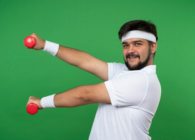 Blij jonge sportieve man met hoofdband en polsband trainen met halters geïsoleerd op groene muur