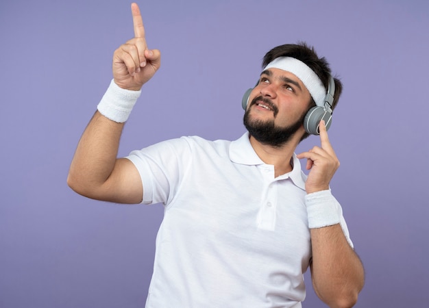 Blij jonge sportieve man opzoeken met hoofdband en polsbandje met koptelefoon wijst naar boven