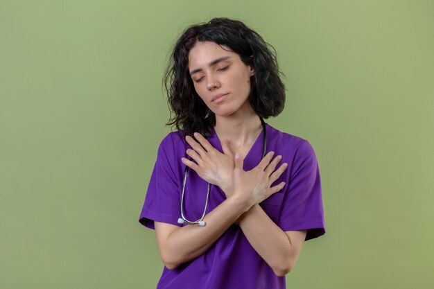 Blij jonge verpleegster die uniform en stethoscoop draagt die zich met handen op borstgebaar bevindt die zich op geïsoleerde groen kruisen die positieve emoties voelen