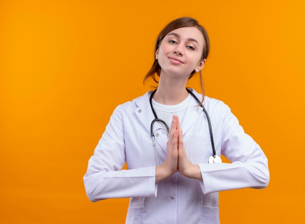 Blij jonge vrouwelijke arts die medische mantel en stethoscoop draagt ?? bidgebaar op geïsoleerde oranje ruimte met exemplaarruimte