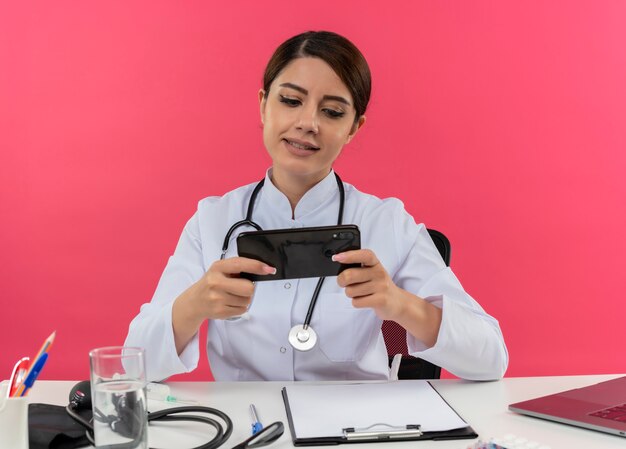 Blij jonge vrouwelijke arts het dragen van medische mantel met stethoscoop zit aan Bureau werken op computer met medische hulpmiddelen speelspel op telefoon met kopie ruimte