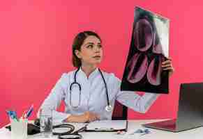 Gratis foto blij jonge vrouwelijke arts medische gewaad dragen met een stethoscoop zit aan bureau werken op computer met medische hulpmiddelen houden en kijken naar x-ray met kopie ruimte
