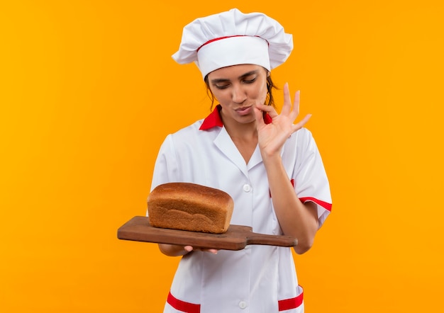Blij jonge vrouwelijke kok die chef-kok uniform draagt en brood op snijplank bekijkt en heerlijk gebaar op geïsoleerde gele muur met exemplaarruimte toont