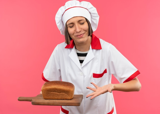 Blij jonge vrouwelijke kok in uniform chef-kok houden en wijzend met de hand op snijplank met brood erop met gesloten ogen geïsoleerd op roze achtergrond