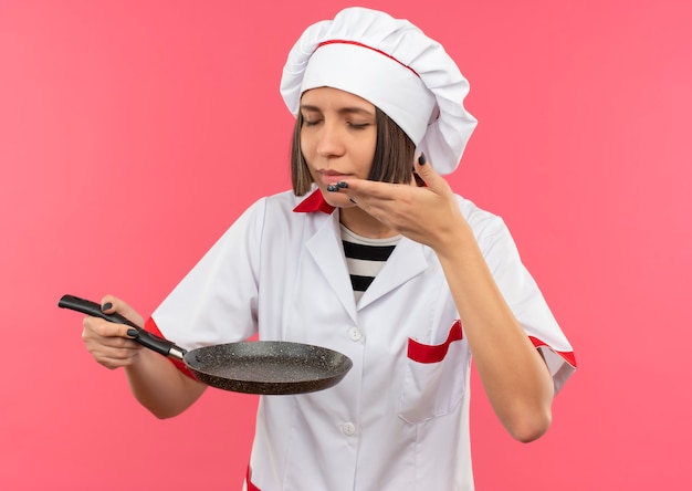 Blij jonge vrouwelijke kok in uniform chef-kok met koekenpan en snuiven met gesloten ogen en hand op lucht houden geïsoleerd op roze muur