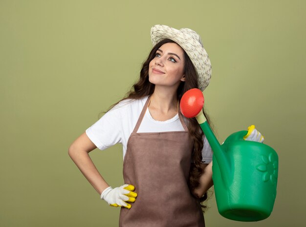 Blij jonge vrouwelijke tuinman in uniform dragen tuinieren hoed en handschoenen houdt gieter en kijkt naar kant geïsoleerd op olijfgroene muur