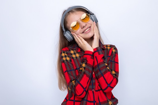 Blij kantelend hoofd handen op wangen leggend mooi klein meisje met rood shirt en bril met koptelefoon