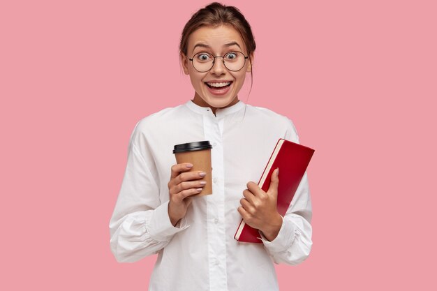 Blij Kaukasisch jong meisje in witte kleren, houdt aromatische afhaalkoffie en boek