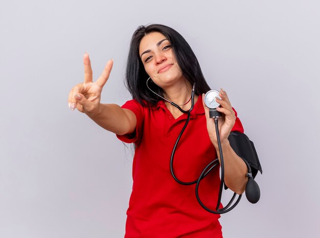 Blij Kaukasische ziek meisje dragen stethoscoop kijken camera haar druk meten met bloeddrukmeter doen vredesteken geïsoleerd op witte achtergrond