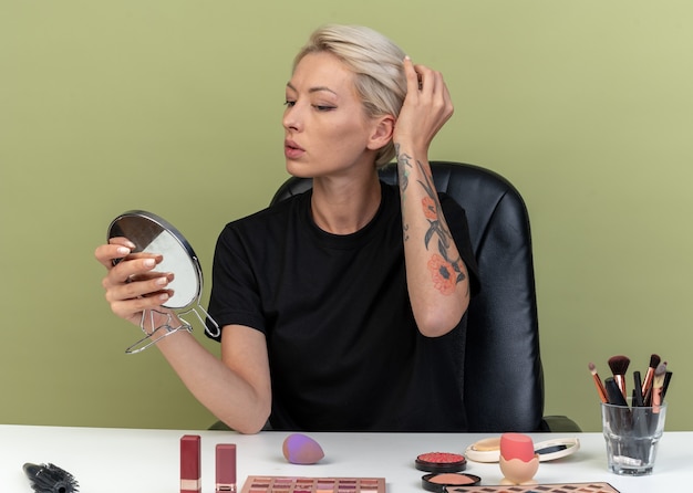 Gratis foto blij kijken naar spiegel, jong mooi meisje zit aan tafel met make-up tools geïsoleerd op olijfgroene muur