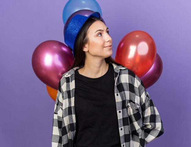 Blij kijkend naar een jonge, mooie vrouw met een feesthoed die voor ballonnen staat geïsoleerd op een blauwe muur