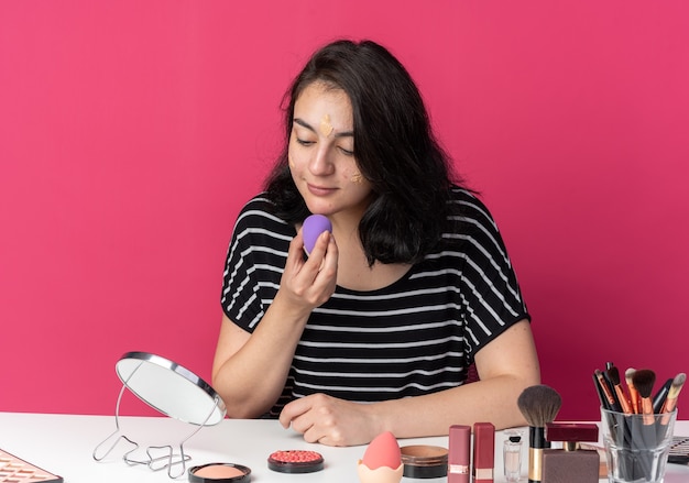 Blij kijkend naar spiegel, jong mooi meisje zit aan tafel met make-uptools die tonuscrème toepast met spons geïsoleerd op roze muur