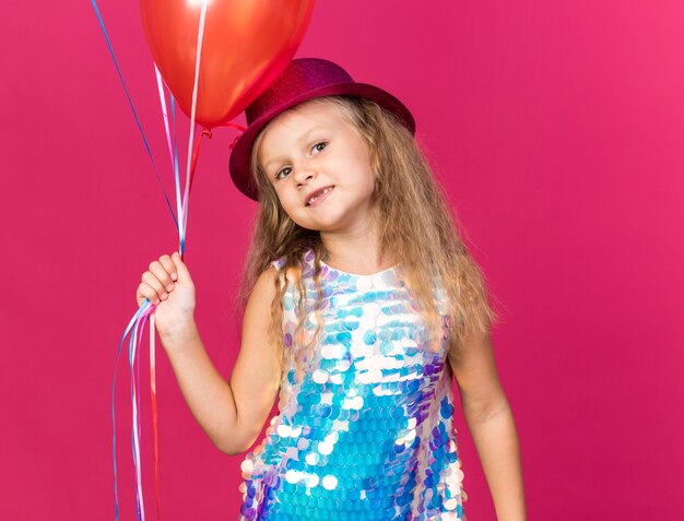 blij klein blond meisje met paarse feestmuts met heliumballonnen geïsoleerd op roze muur met kopieerruimte