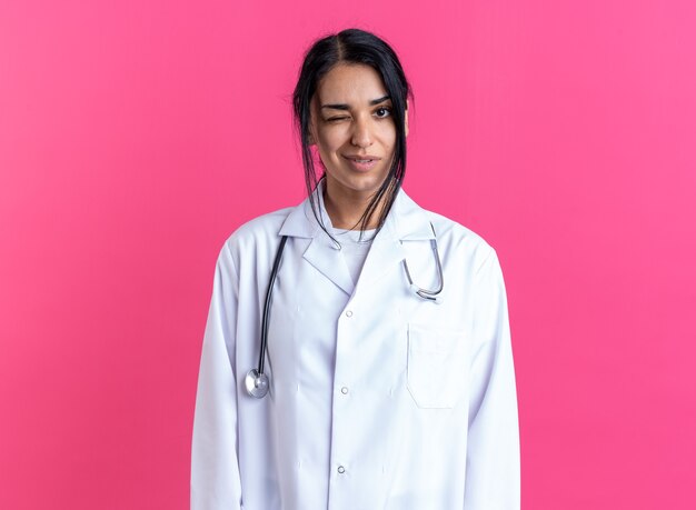Blij knipperde jonge vrouwelijke arts die medische mantel draagt met stethoscoop geïsoleerd op roze muur
