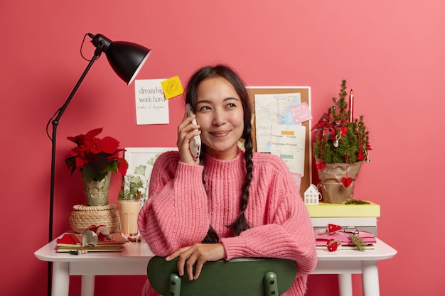 Blij Koreaans meisje luistert goed nieuws op smartphone, kijkt weg, nonchalant gekleed, poseert in eigen huiskast, bereidt zich voor op sessie, kleine versierde kerstboom op tafel